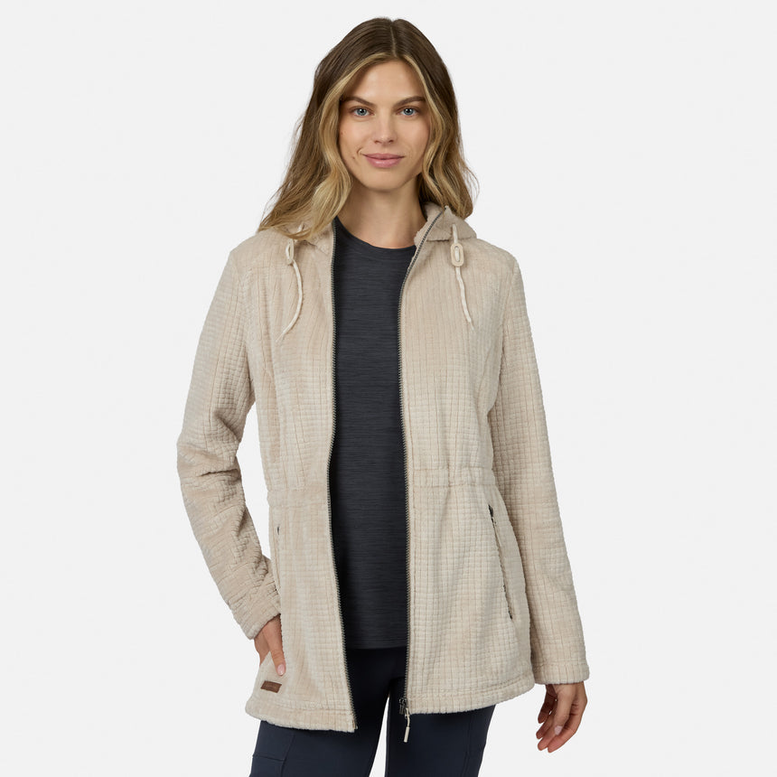 A woman models the Free Country Women’s Grid Butter Pile® Anorak Jacket in light beige, hooded with drawstrings, over a dark shirt and pants, standing with one hand in her pocket against a plain white background.