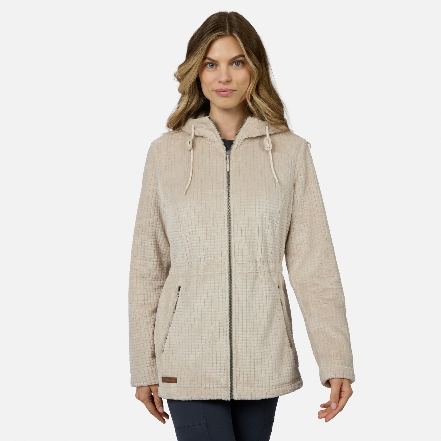 A woman with long, wavy hair wears the Free Country Women’s Grid Butter Pile® Anorak Jacket in light beige, featuring drawstrings, standing against a plain white background.