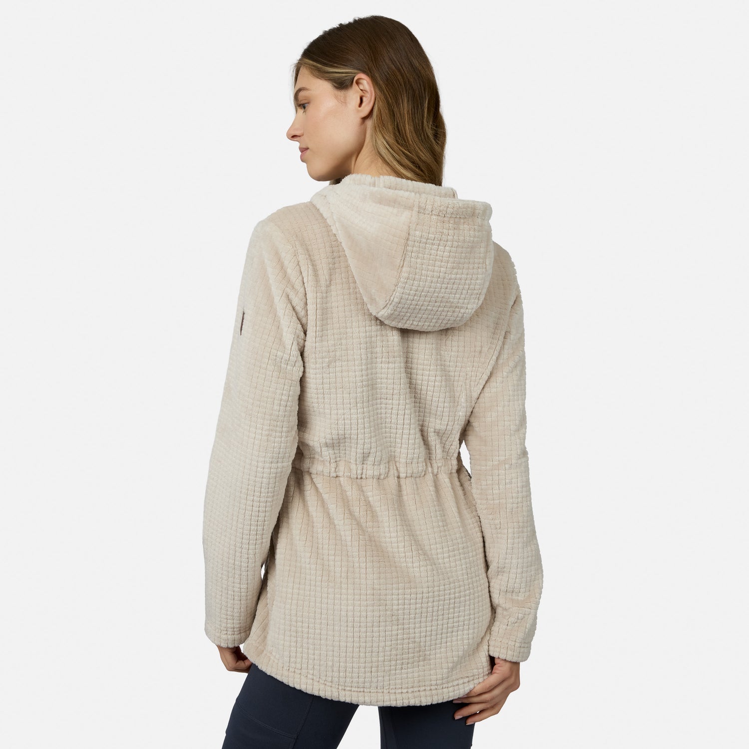 A woman with light brown hair models the Free Country Women’s Grid Butter Pile® Anorak Jacket in beige with dark pants, standing with her back to the camera against a plain white background.
