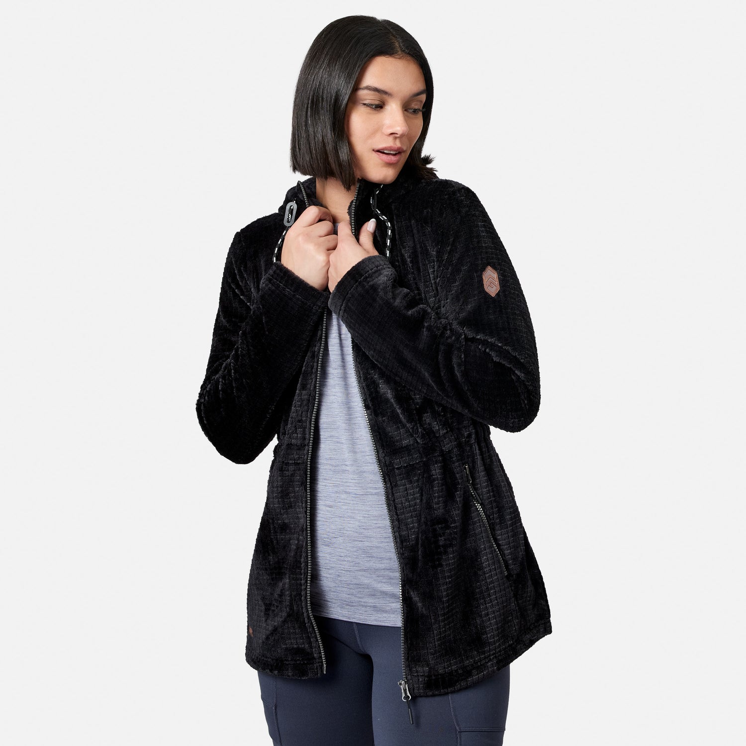 A woman stands against a plain white background, looking down and holding the collar of her Free Country Women’s Grid Butter Pile® Anorak Jacket, worn over a light gray top and dark pants.