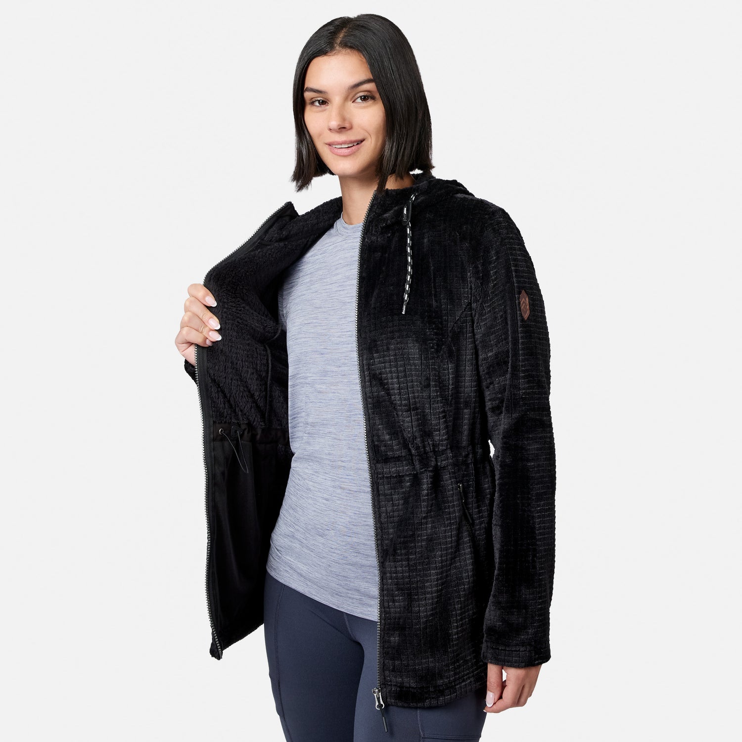 A person wearing a gray shirt and navy pants smiles at the camera against a plain white background while opening the long, black, textured Free Country Women’s Grid Butter Pile® Anorak Jacket to reveal its soft interior.