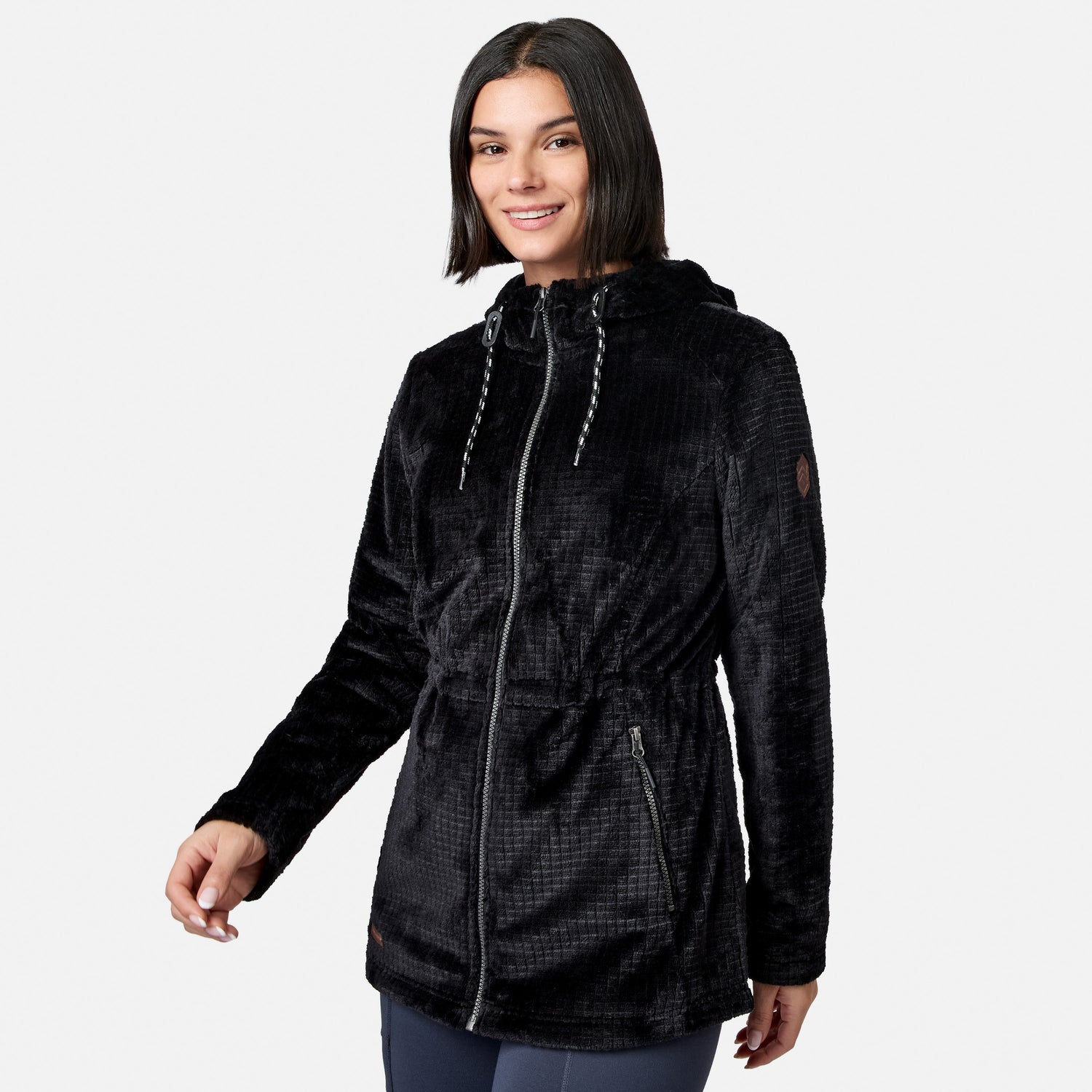 A woman wearing the Free Country Women’s Grid Butter Pile® Anorak Jacket, featuring a black textured design with a hood and drawstring, smiles while standing against a plain white background.