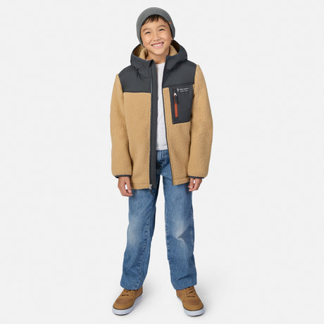 A young boy wearing a tan and dark gray fleece jacket, blue jeans, brown shoes, and a gray beanie, standing against a light gray studio background.
