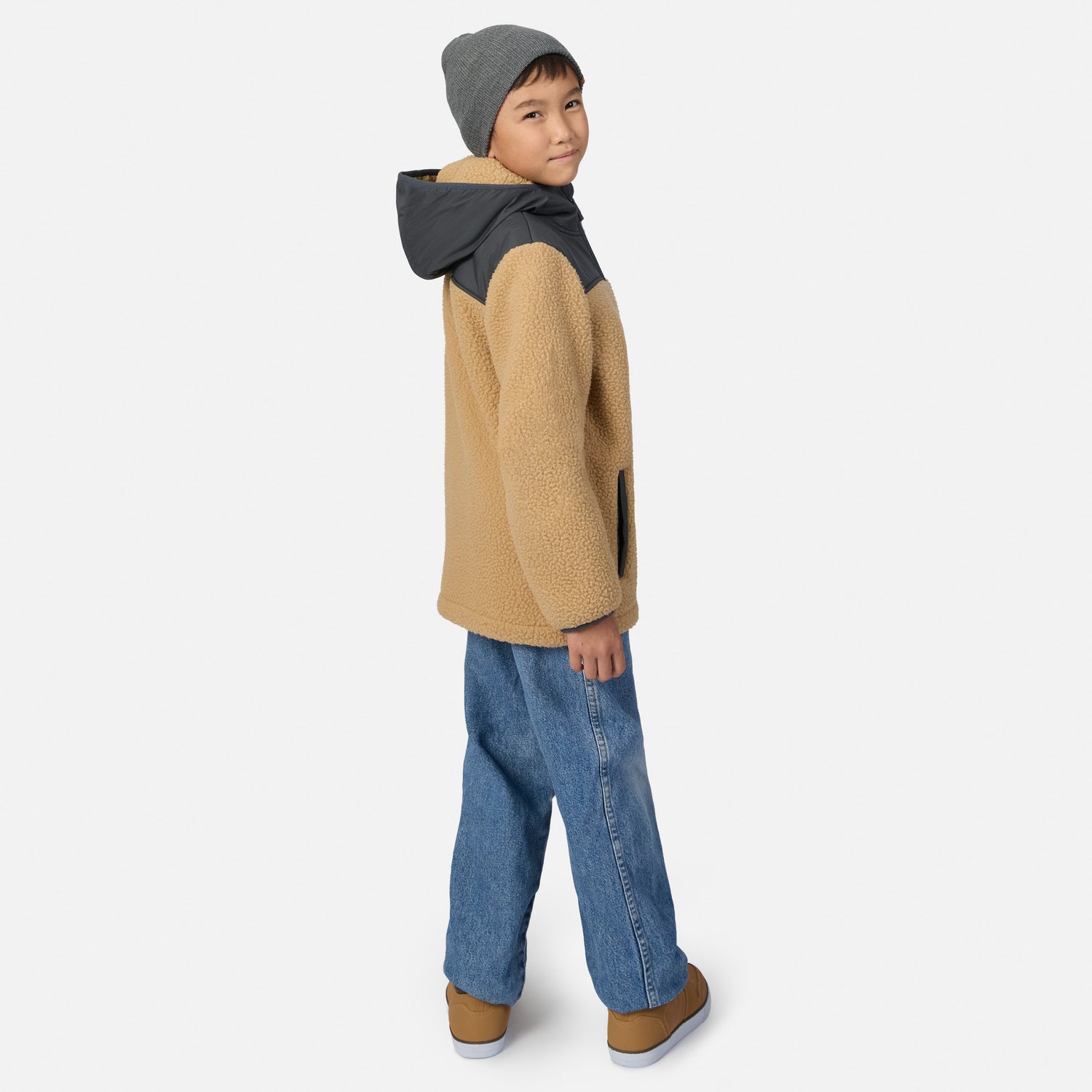 A child model wearing a tan sherpa fleece hooded jacket with dark gray shoulder panels, blue jeans, and a gray beanie, shown from a side-back angle against a light gray studio background.