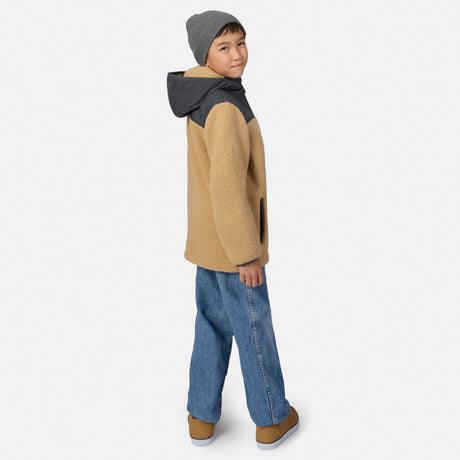 A child model wearing a tan sherpa fleece hooded jacket with dark gray shoulder panels, blue jeans, and a gray beanie, shown from a side-back angle against a light gray studio background.