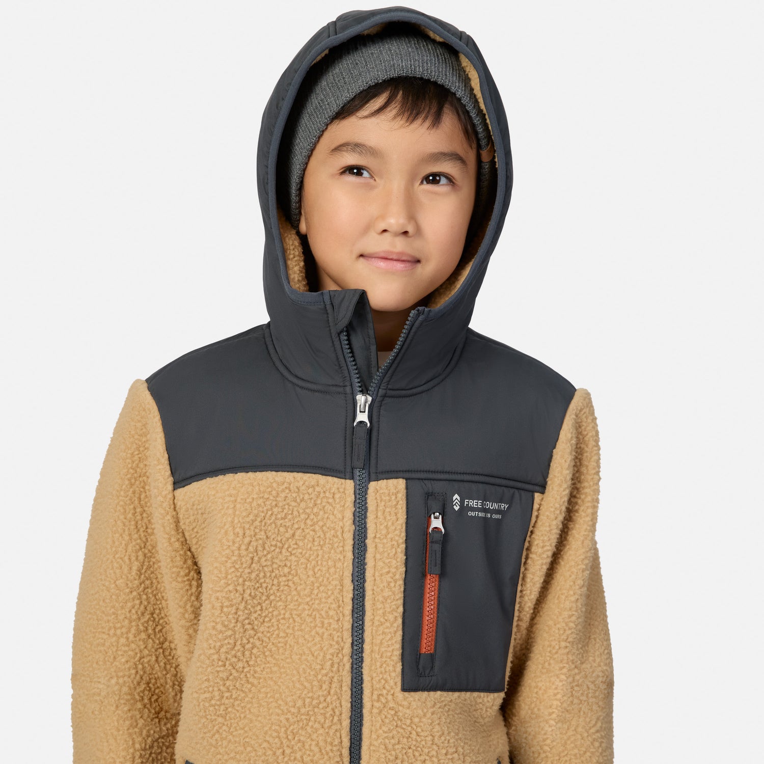 A child wearing a tan and dark gray fleece zip-up jacket with a hood, paired with a gray beanie, against a light gray background.