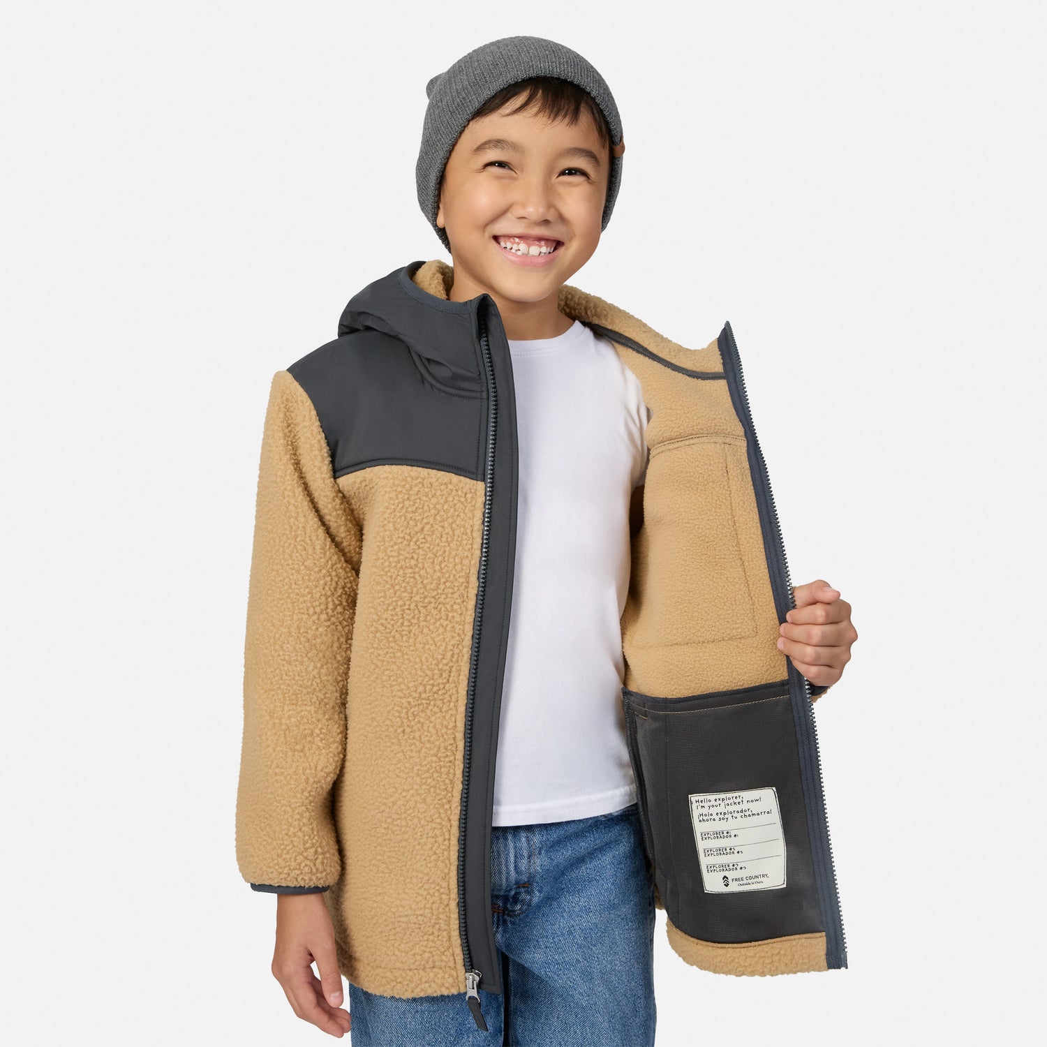 A smiling young boy wearing a tan and dark gray fleece zip-up jacket, a gray beanie, a white t-shirt, and blue jeans, holding the jacket open to show the interior pocket against a light gray background.