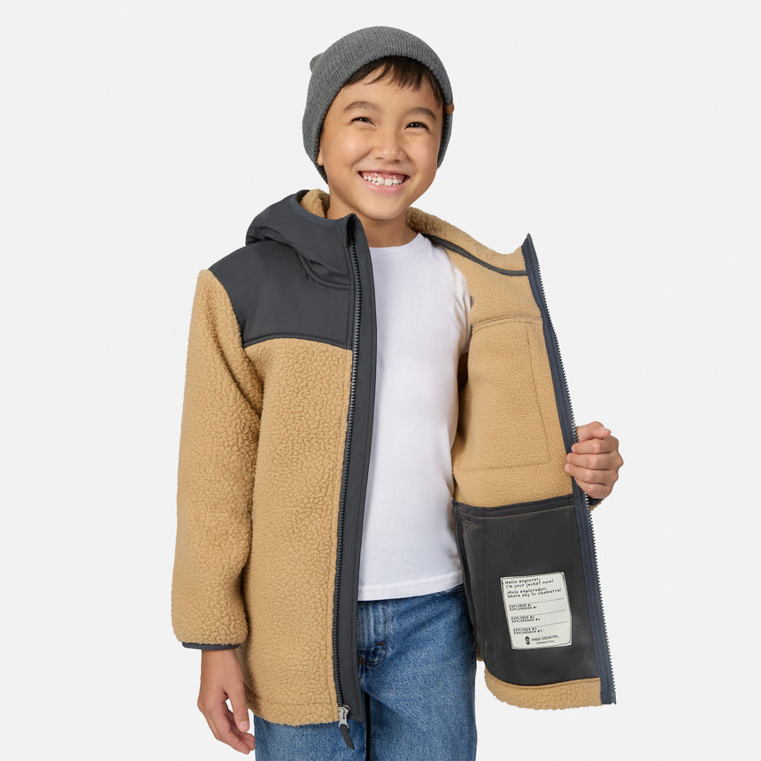 A smiling young boy wearing a tan and dark gray fleece zip-up jacket, a gray beanie, a white t-shirt, and blue jeans, holding the jacket open to show the interior pocket against a light gray background.