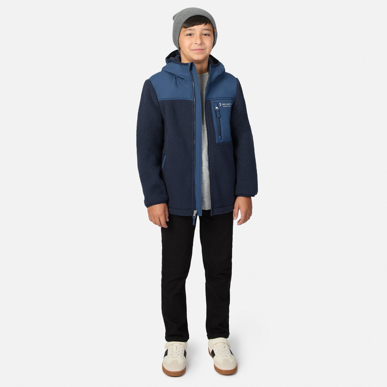 A child wearing a navy and blue fleece jacket, a gray beanie, a light gray shirt, black pants, and off-white sneakers, standing against a light gray studio background.
