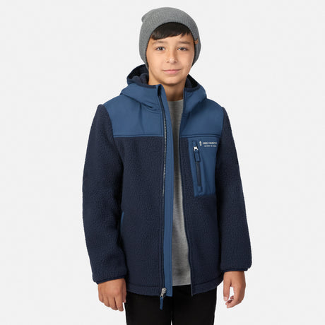 A child wearing a navy and blue fleece jacket with a gray beanie and gray t-shirt, standing against a light gray background.