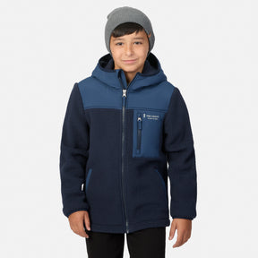 A child wearing a navy and blue fleece zip-up jacket with a hood, paired with a gray beanie, standing against a light gray background.