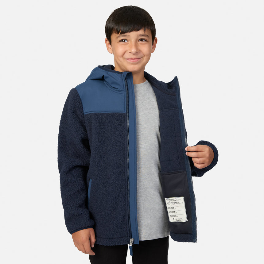 A young boy wearing a navy blue and slate blue fleece hooded jacket over a gray t-shirt, standing against a light gray studio background.