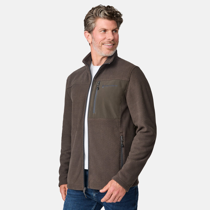 [Kolby is 6'2" wearing size M.] Men’s Free Country brown textured full-zip jacket with high collar and zippered chest pocket in contrast fabric—featuring a streamlined silhouette for cool-weather utility. Paired with blue jeans for a classic, casual outdoor look.
