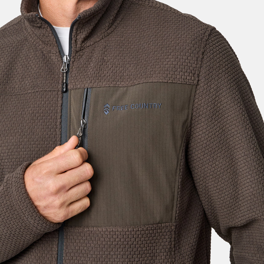 [Kolby is 6'2" wearing size M.] Men’s Free Country brown textured full-zip jacket with high collar and zippered chest pocket in contrast fabric—featuring a streamlined silhouette for cool-weather utility. Paired with blue jeans for a classic, casual outdoor look.