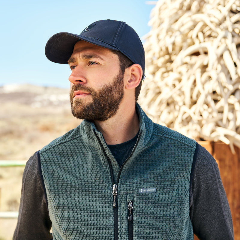 Man wearing a black cap and green Free Country vest standing outdoors with a blurred natural background