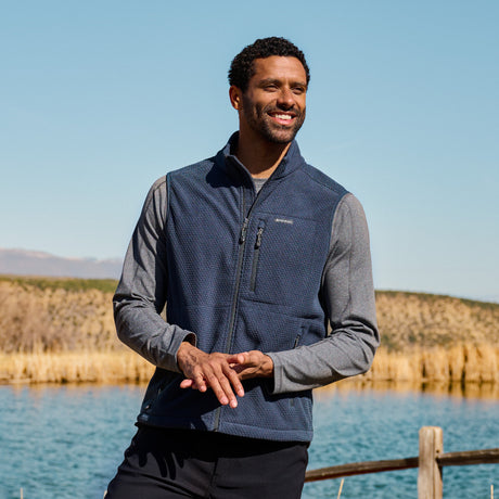 Man wearing a blue Free Country vest standing by a lake with mountains in the background