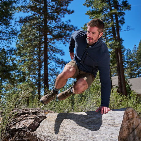 Wearing the Free Country Men’s Calabaza II Brick Fleece Jacket and matching shorts, a man leaps over a large fallen tree trunk in a forest of towering pine trees beneath a clear blue sky, highlighting the lightweight fleece jacket’s exceptional comfort during his outdoor adventure.