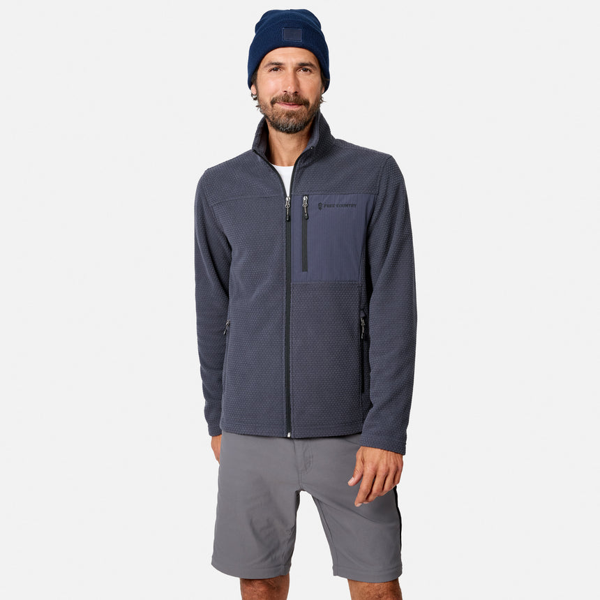 A man wears a navy beanie, storm-colored Free Country Men’s Calabaza II Brick Fleece Jacket, and gray shorts while facing the camera against a plain white background.