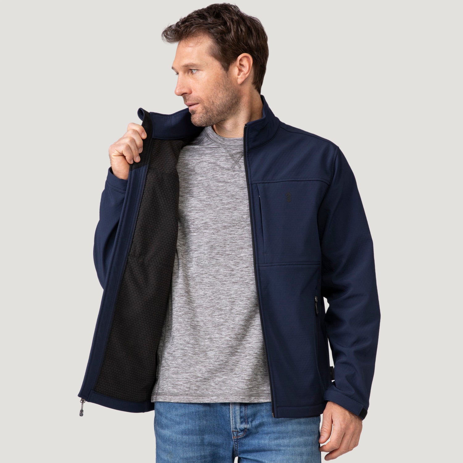 A man wears a dark blue Men’s Stretch Super Softshell® Jacket over a gray t-shirt, with the jacket partially open to reveal a black interior lining.
