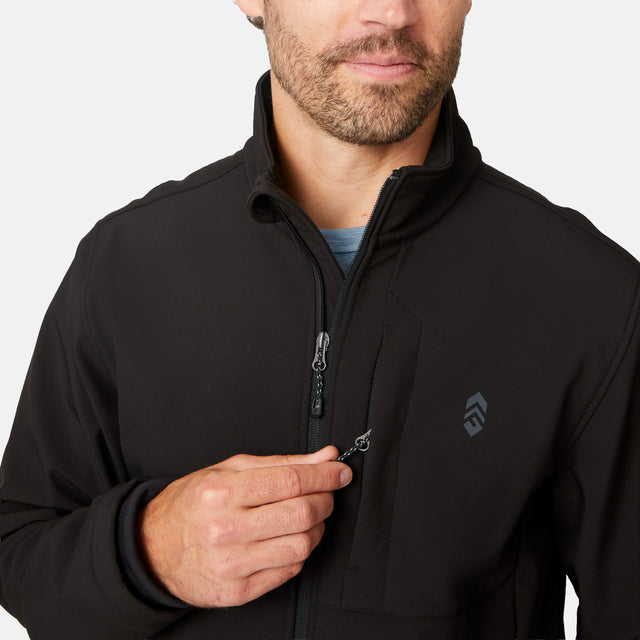 [Matt is 6'2" wearing size M.] Men’s Free Country black zip-front softshell jacket with high stand collar, subtle chest logo, and minimalistic design—featuring streamlined silhouette and long sleeves. Worn with blue pants for a clean, versatile look suited to cool-weather outings.