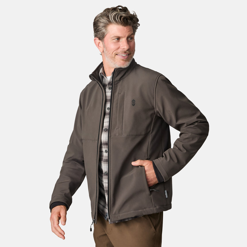 [Kolby is 6'2" wearing size M.] Men’s Free Country brown zip-front softshell jacket with high stand collar, subtle chest logo, and minimalistic design—featuring streamlined silhouette and long sleeves. Worn with blue pants for a clean, versatile look suited to cool-weather outings.