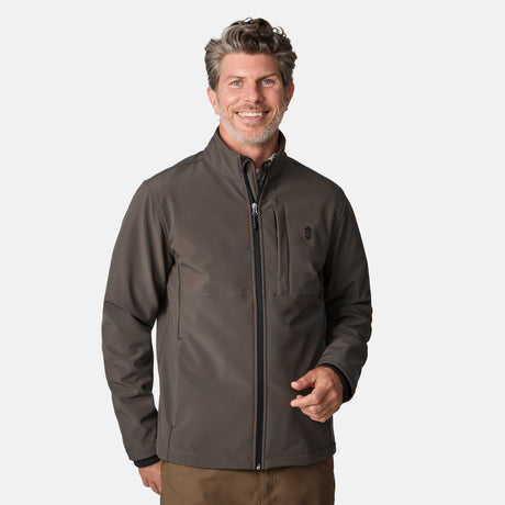 [Kolby is 6'2" wearing size M.] Men’s Free Country brown zip-front softshell jacket with high stand collar, subtle chest logo, and minimalistic design—featuring streamlined silhouette and long sleeves. Worn with blue pants for a clean, versatile look suited to cool-weather outings.