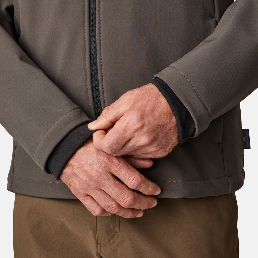 [Kolby is 6'2" wearing size M.] Men’s Free Country brown zip-front softshell jacket with high stand collar, subtle chest logo, and minimalistic design—featuring streamlined silhouette and long sleeves. Worn with blue pants for a clean, versatile look suited to cool-weather outings.