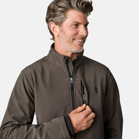 [Kolby is 6'2" wearing size M.] Men’s Free Country brown zip-front softshell jacket with high stand collar, subtle chest logo, and minimalistic design—featuring streamlined silhouette and long sleeves. Worn with blue pants for a clean, versatile look suited to cool-weather outings.