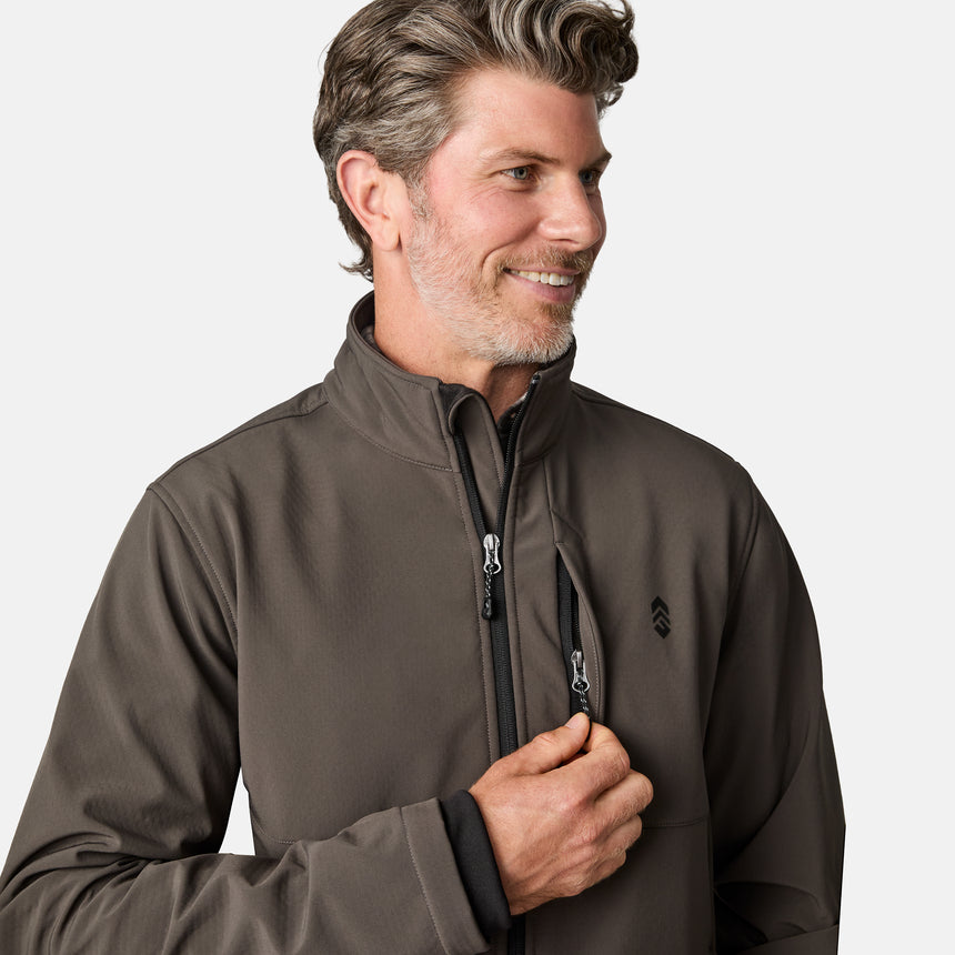 [Kolby is 6'2" wearing size M.] Men’s Free Country brown zip-front softshell jacket with high stand collar, subtle chest logo, and minimalistic design—featuring streamlined silhouette and long sleeves. Worn with blue pants for a clean, versatile look suited to cool-weather outings.