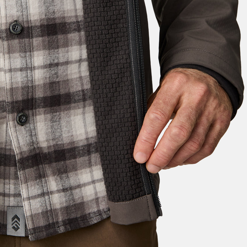 [Kolby is 6'2" wearing size M.] Men’s Free Country brown zip-front softshell jacket with high stand collar, subtle chest logo, and minimalistic design—featuring streamlined silhouette and long sleeves. Worn with blue pants for a clean, versatile look suited to cool-weather outings.