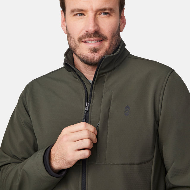 [Joe is 6'0" wearing size M.] Men’s Free Country olive green zip-front softshell jacket with high stand collar, subtle chest logo, and minimalistic design—featuring streamlined silhouette and long sleeves. Worn with blue pants for a clean, versatile look suited to cool-weather outings.