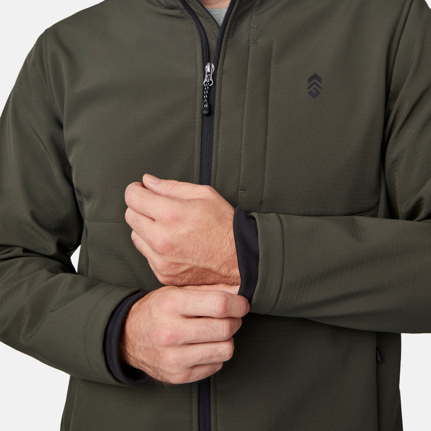 [Joe is 6'0" wearing size M.] Men’s Free Country olive green zip-front softshell jacket with high stand collar, subtle chest logo, and minimalistic design—featuring streamlined silhouette and long sleeves. Worn with blue pants for a clean, versatile look suited to cool-weather outings.