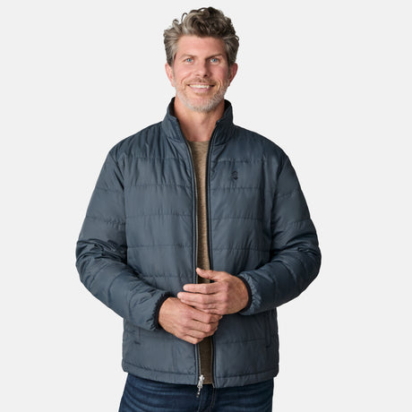 A bearded man with short, wavy hair smiles while wearing the Free Country Men's Atalaya III 3-in-1 Systems Jacket—blue, water resistant, and made from recycled REPREVE—over a tan shirt and jeans against a plain white background.