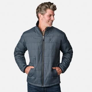 A man with short, wavy hair smiles and looks to the side, casually dressed in jeans and wearing the Free Country Men's Atalaya III 3-in-1 Systems Jacket in gray. His hands are in his pockets as he stands against a plain white background.