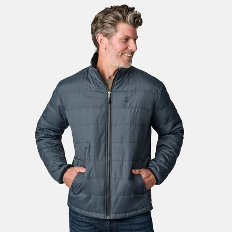 A man with short, wavy hair smiles and looks to the side, casually dressed in jeans and wearing the Free Country Men's Atalaya III 3-in-1 Systems Jacket in gray. His hands are in his pockets as he stands against a plain white background.