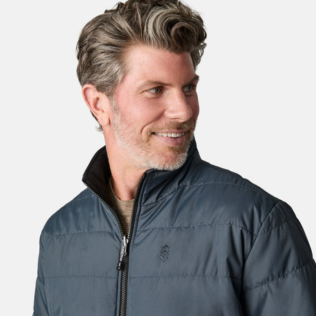 A man with wavy, gray-streaked hair and a beard is wearing the Free Country Men's Atalaya III 3-in-1 Systems Jacket over a gray shirt. He is smiling and looking to his left against a plain light background.