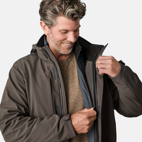 A smiling man with wavy brown hair and a beard zips up the Free Country Men's Atalaya III 3-in-1 Systems Jacket, made from recycled REPREVE fabric, over a beige shirt against a plain light background.