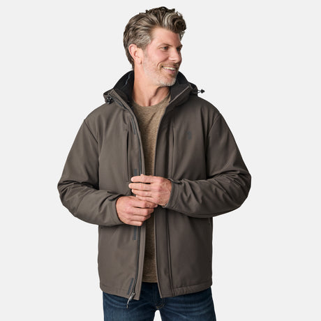 A man wearing the Free Country Men's Atalaya III 3-in-1 Systems Jacket in gray over a tan shirt stands against a white background, smiling and looking left with his hands near the jacket zipper.