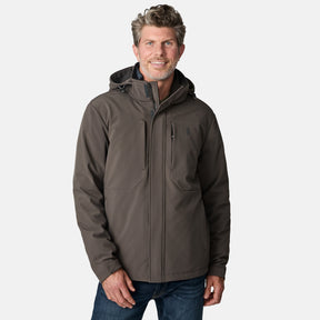 A man with short, wavy hair is wearing the Free Country Men's Atalaya III 3-in-1 Systems Jacket in dark gray and blue jeans, standing and smiling against a plain light gray background.