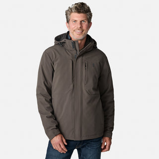 A man with short, wavy hair is wearing the Free Country Men's Atalaya III 3-in-1 Systems Jacket in dark gray and blue jeans, standing and smiling against a plain light gray background.