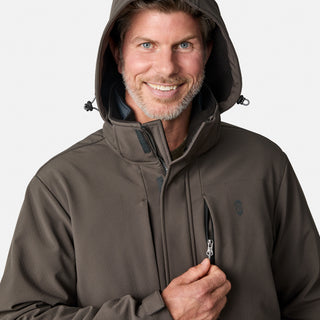 A man with a trimmed beard and mustache smiles as he unzips a chest pocket on the Free Country Men's Atalaya III 3-in-1 Systems Jacket in brown, wearing the hood up against a plain background.