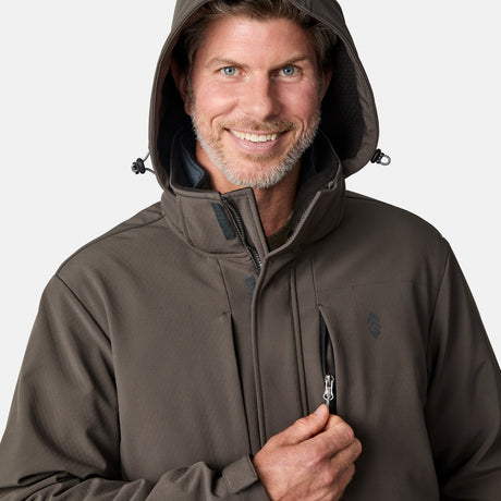A man with a trimmed beard and mustache smiles as he unzips a chest pocket on the Free Country Men's Atalaya III 3-in-1 Systems Jacket in brown, wearing the hood up against a plain background.