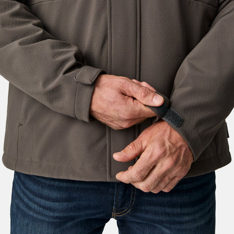 A person wearing the Free Country Men's Atalaya III 3-in-1 Systems Jacket in brown and blue jeans adjusts the Velcro strap on the jacket's sleeve. Only their torso and hands are visible.