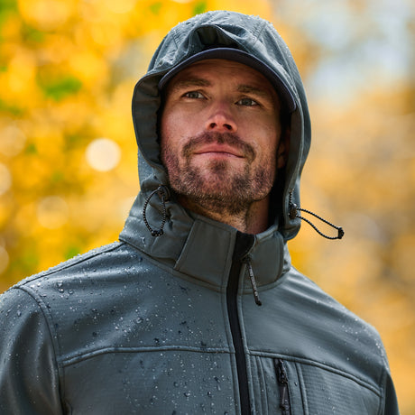 A man wearing a dark gray waterproof hooded jacket with water droplets on it, set against a blurred background of golden autumn trees.