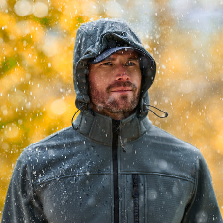 A man wearing a dark gray hooded waterproof jacket stands outdoors in falling snow with a blurred golden-yellow background.