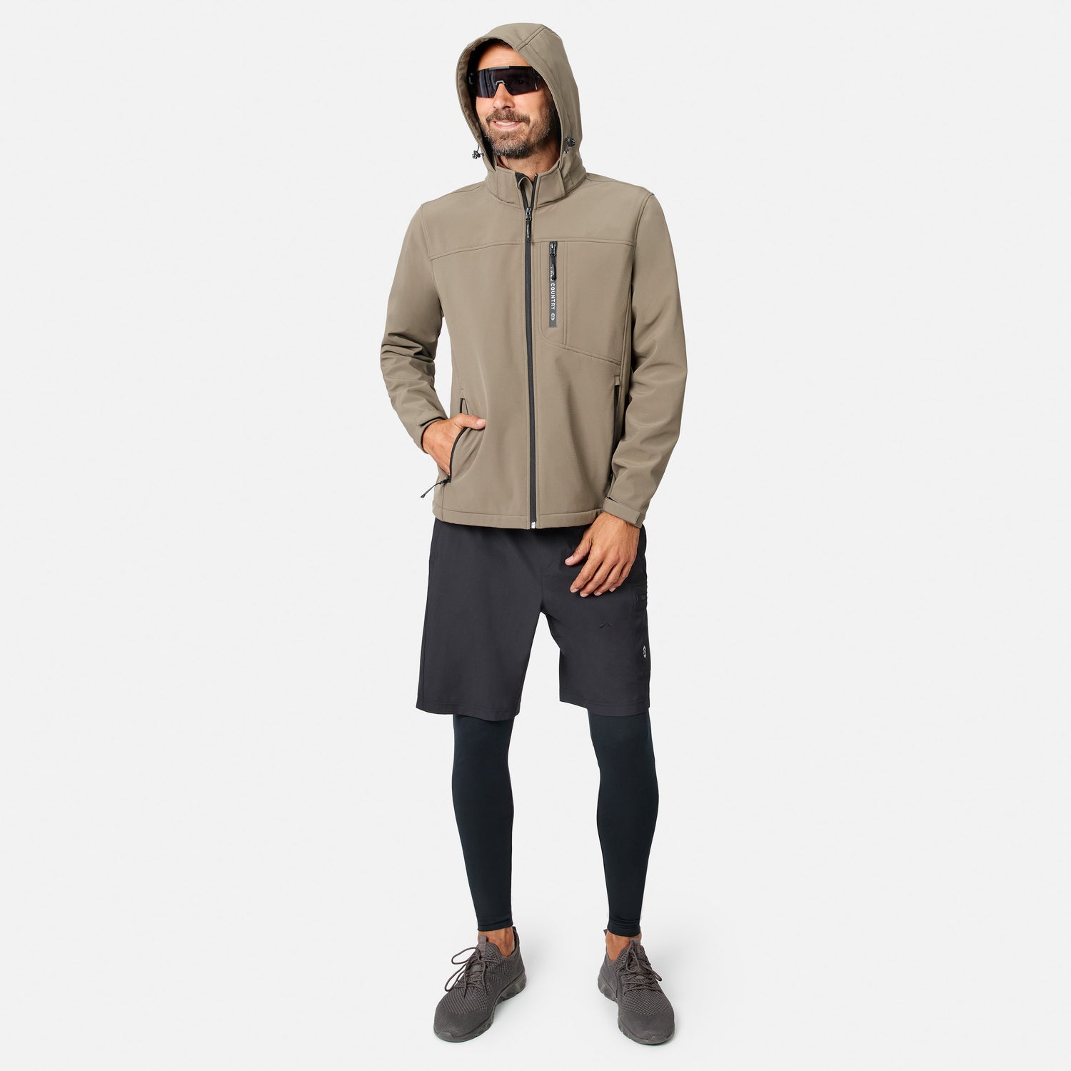 [Model is wearing size M.] A man wearing a light brown hooded zip-up jacket, black athletic shorts, black leggings, and gray sneakers, standing against a light gray background.