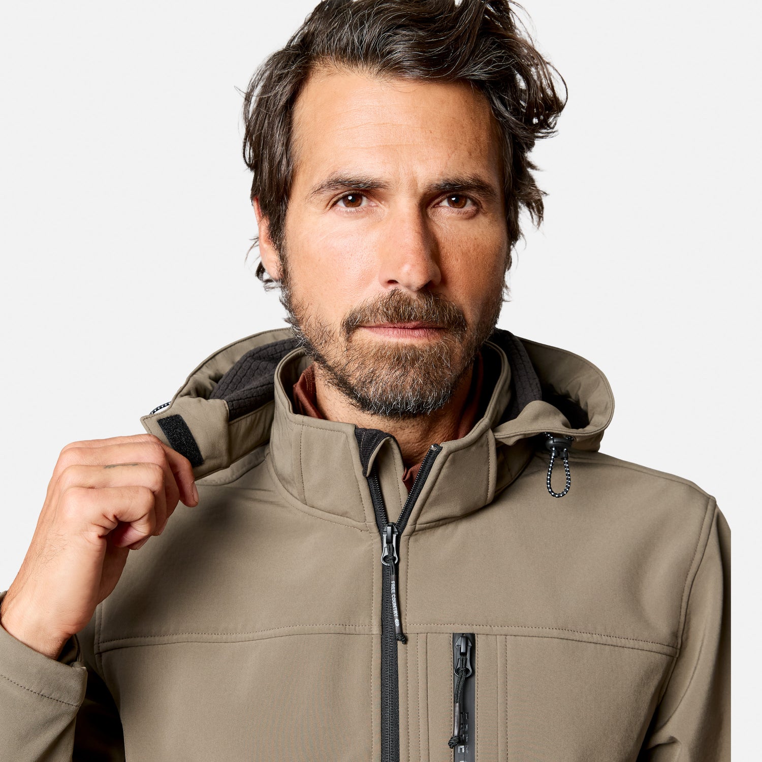 [Model is wearing size M.] A man wearing a light brown softshell hooded jacket, shown from the chest up against a light gray studio background. He is adjusting the collar with his right hand.