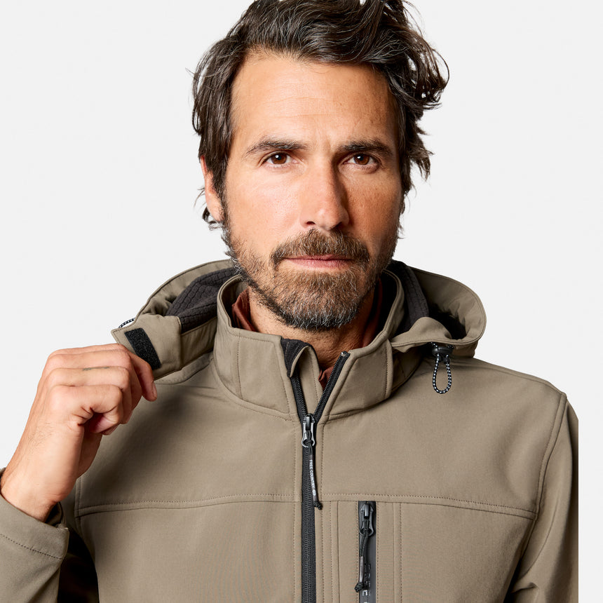 [Model is wearing size M.] A man wearing a light brown softshell hooded jacket, shown from the chest up against a light gray studio background. He is adjusting the collar with his right hand.