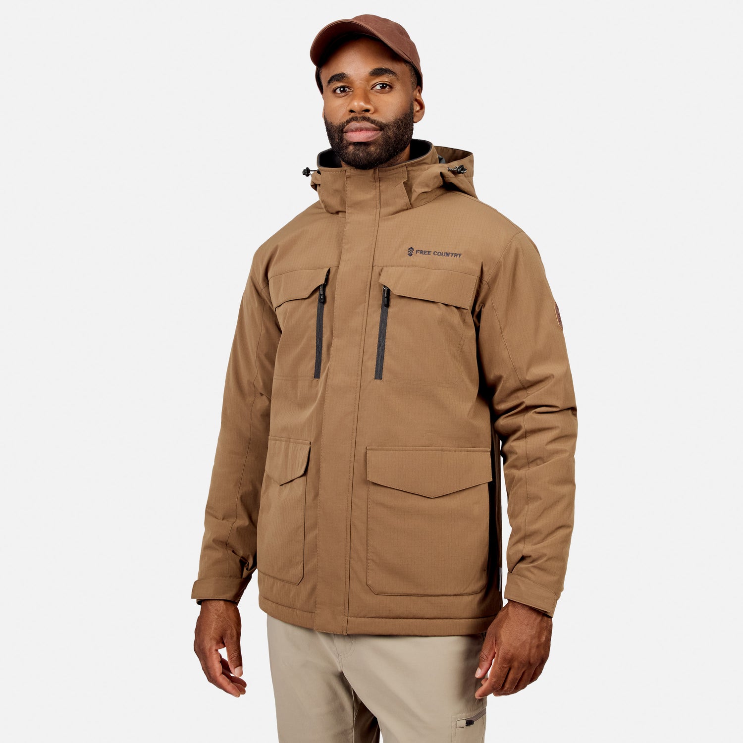 [Model is wearing size M.] A man wearing a tan hooded outdoor jacket with multiple zippered and flap pockets, paired with light khaki pants and a brown cap, photographed against a light gray studio background.