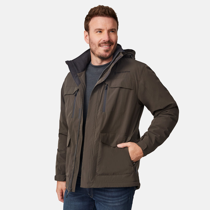 [Joe is 6'0" wearing size M.] Men’s Free Country brown hooded utility jacket featuring dual zippered chest pockets, flap waist pockets, and rugged outdoor styling—paired with blue jeans for a functional, adventure-ready look.
